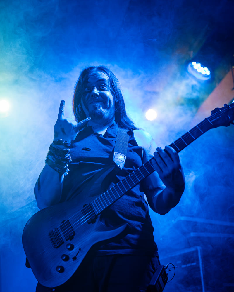 Riese auf der Bühne in blauem Licht, mit der Gitarre, die freie Hand zur Pommesgabel erhoben