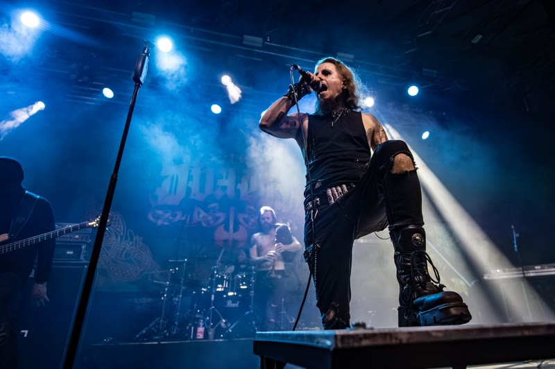 Simon singt mit einem Fuß auf dem Monitor, dahinter die Band vor dem Banner
